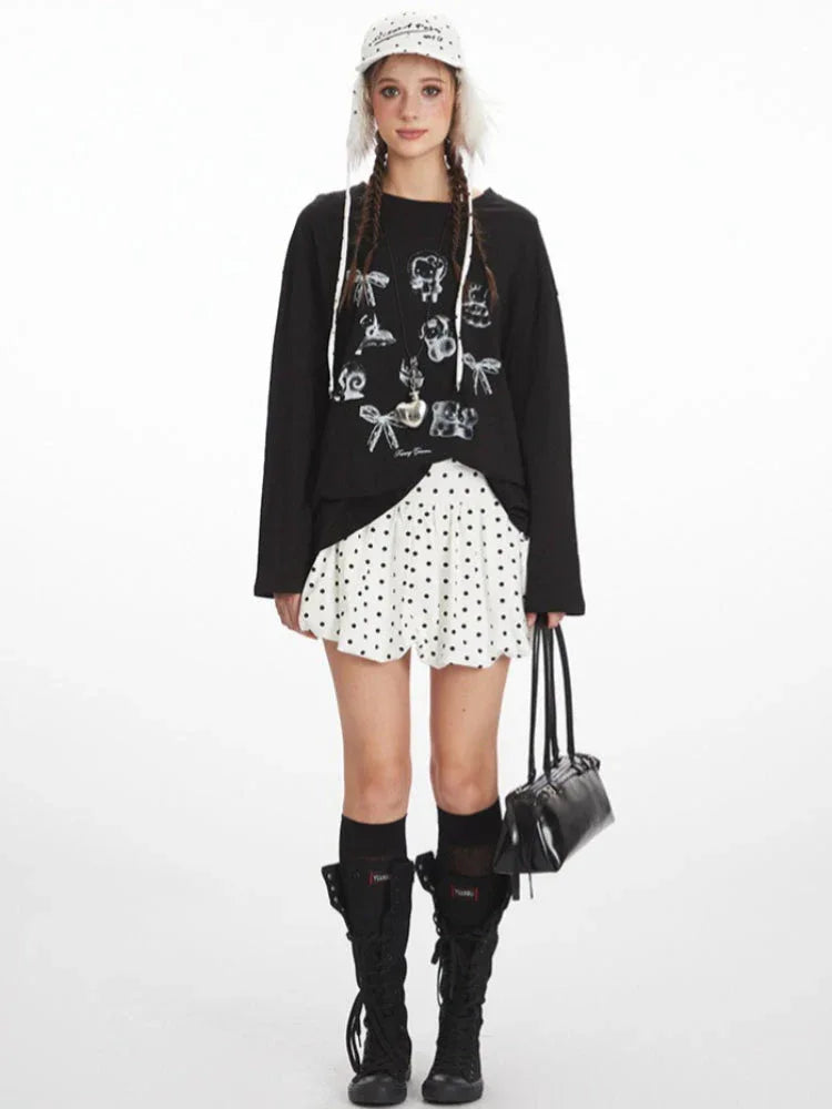 Full length studio shot of a young woman wearing a black long-sleeved graphic t-shirt, a white polka dot mini skirt, black knee-high socks, and black boots. She is also wearing a white polka dot bucket hat with fur trim and carrying a black handbag.