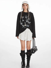 Full length studio shot of a young woman wearing a black long-sleeved graphic t-shirt, a white polka dot mini skirt, black knee-high socks, and black boots. She is also wearing a white polka dot bucket hat with fur trim and carrying a black handbag.