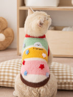 A cream-colored cat wearing a colorful striped sweater with embroidered details and appliqued stars sits facing away from the camera.