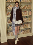 A young woman leans against a bookshelf wearing a brown sweatshirt, grey cycling shorts, and a blue and white plaid scarf.