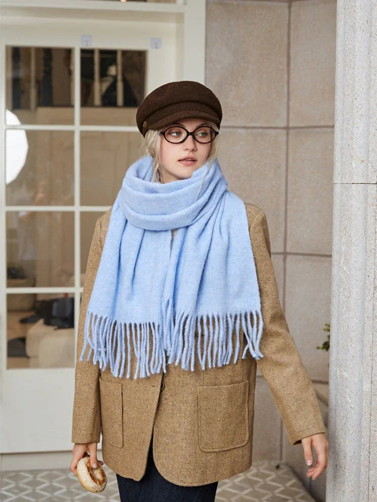 A young woman wearing a light blue fringed scarf and a brown tweed jacket stands outside a building.