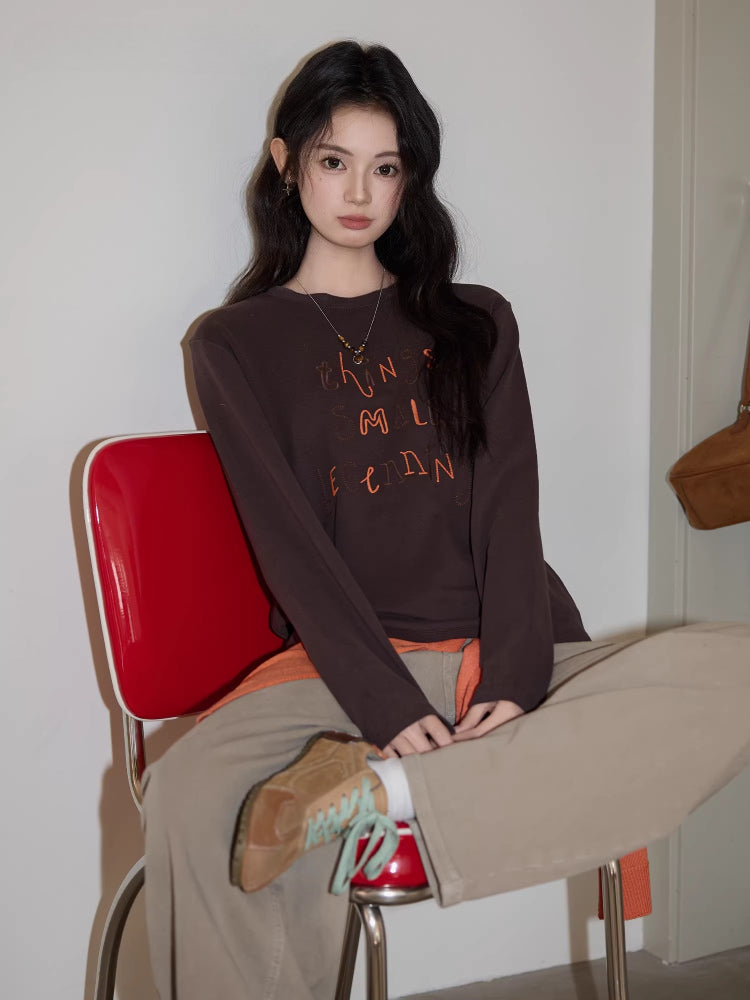 Young woman wearing a brown long-sleeved graphic t-shirt and beige pants, sitting on a red chair