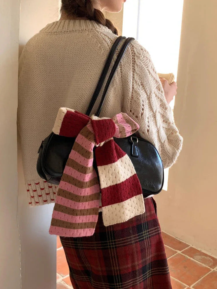 A close-up of a black handbag with a striped scarf tied around the handle, as worn by a person wearing a cream cable knit sweater and plaid skirt.