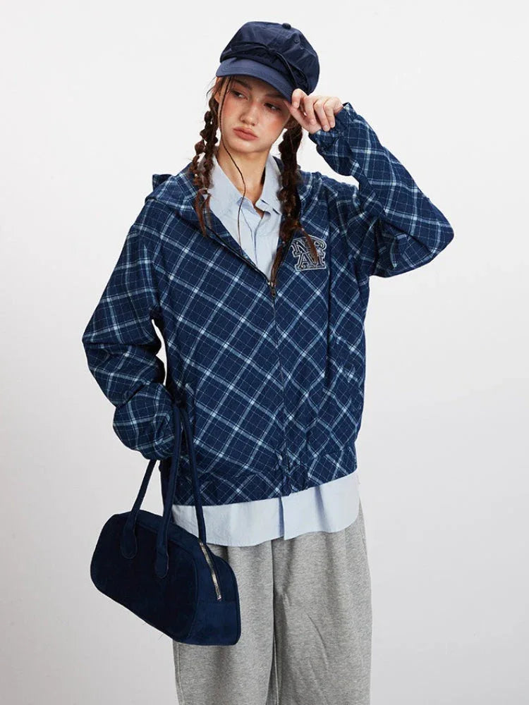 Close-up of a model wearing a blue plaid hooded jacket, light blue collared shirt, and a navy blue cap, adjusting the hood.