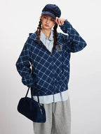 Close-up of a model wearing a blue plaid hooded jacket, light blue collared shirt, and a navy blue cap, adjusting the hood.