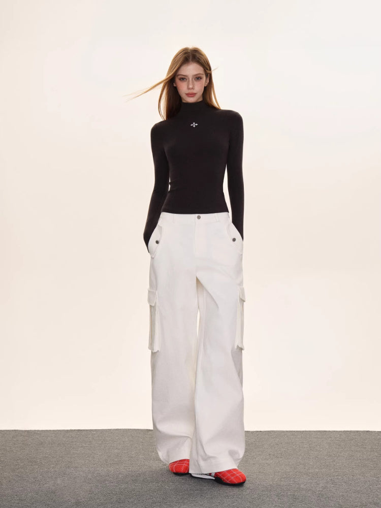 Full-body shot of a woman in a black turtleneck top with a white embroidered cross, and wide-leg white cargo pants.