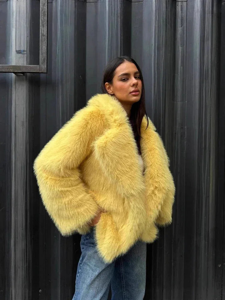 Close-up of a woman wearing a yellow faux fur jacket and blue jeans, with her hand in her pocket, against a corrugated metal wall.