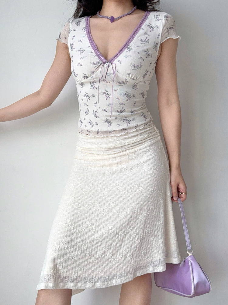 Full body shot of a woman wearing a white floral print top, white textured skirt, and a purple handbag.