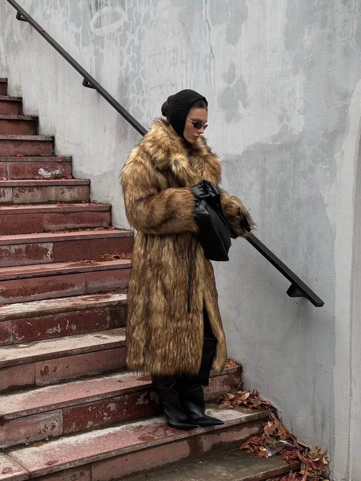 Woman wearing a long brown faux fur coat with a black scarf, black boots, and a black bag, looking down on stairs.