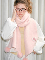 Woman wearing a pink fuzzy knit scarf with pom-pom embellishments, touching her forehead.