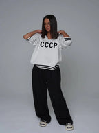Woman wearing a white CCCP knit sweater with black stripes on the cuffs and hem, and black wide-leg pants.