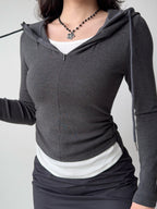 Side view of a woman wearing a dark grey hooded zip-up top, white tank, and black skirt, with a handbag.