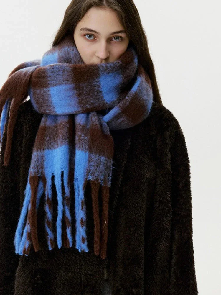 A woman models a cozy, oversized blue and brown plaid scarf with fringe, wrapped around her neck and covering her mouth.