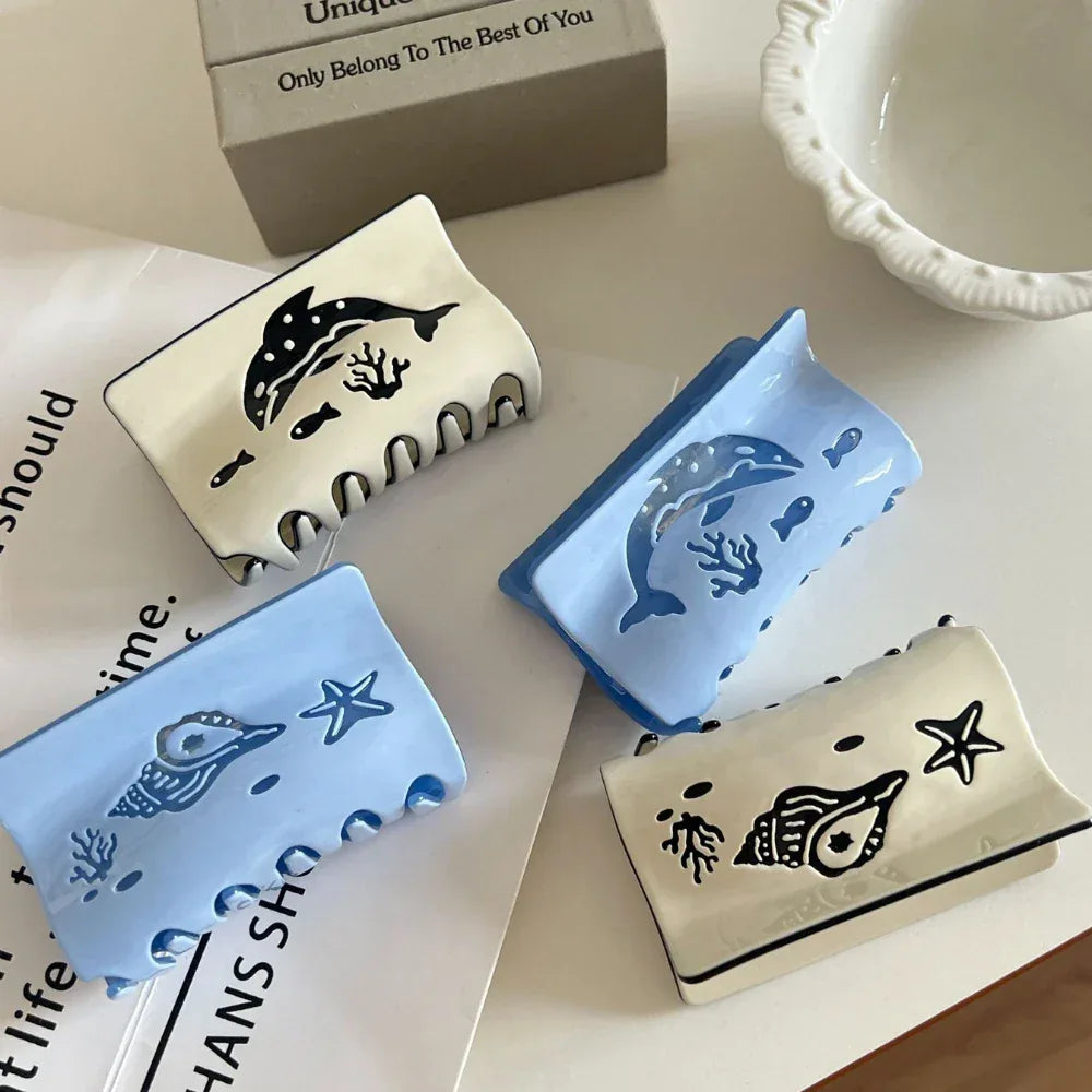 Four ocean-themed hair claw clips arranged on a table, two with dolphin designs and two with seashell designs