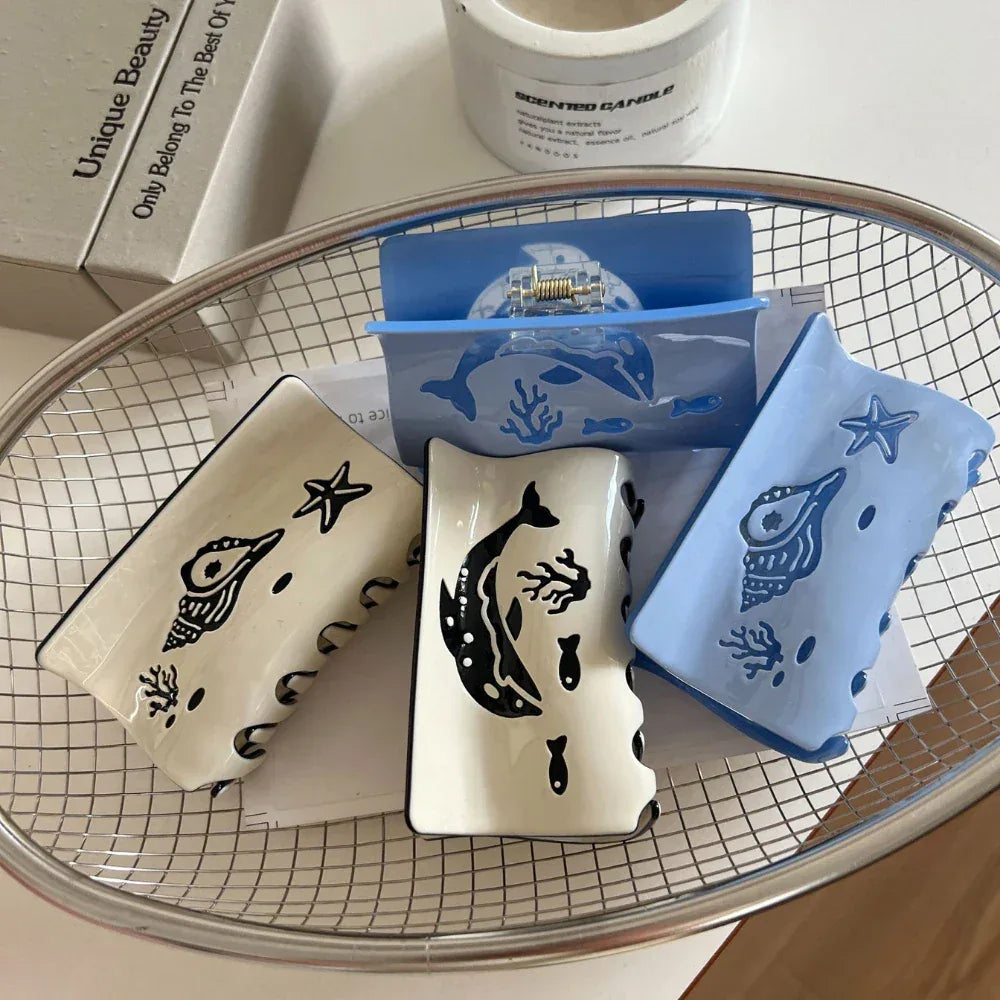 Four hair claw clips with marine motifs, including dolphins and seashells, displayed in a metal basket