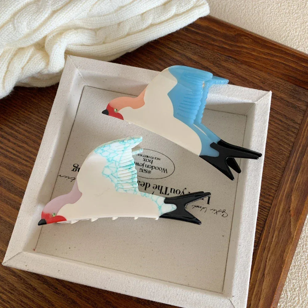 Two bird-shaped hair clips, one with blue wings and the other with blue-veined white wings, placed in a white box.