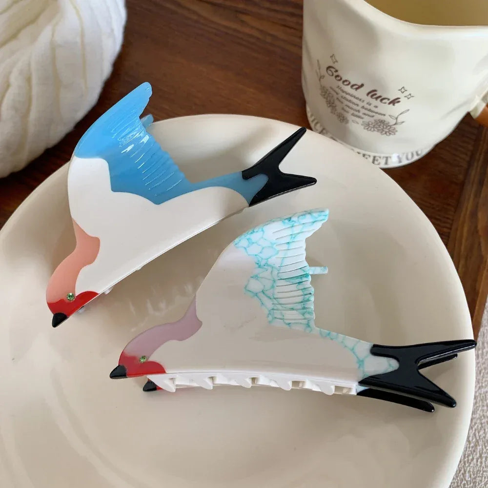 Two bird-shaped hair clips, one with blue wings and the other with blue-veined white wings, arranged on a white plate.
