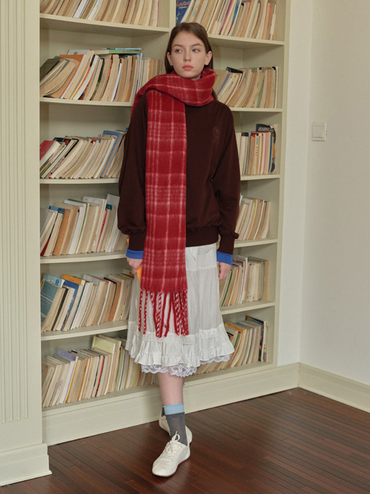 A young woman stands in front of a bookshelf wearing a brown sweatshirt, a white ruffled skirt, and a red plaid scarf, holding a book.
