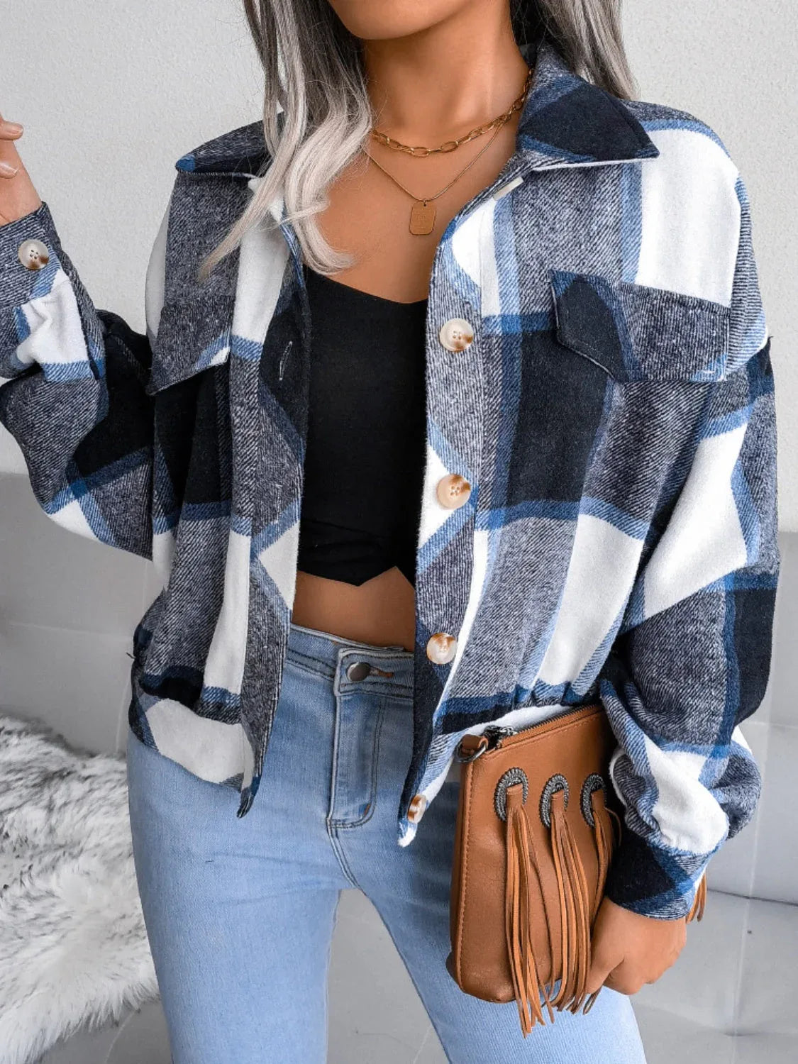 Woman wearing a blue, white, and black plaid flannel shirt jacket over a black crop top and light wash jeans, holding a brown fringed clutch.