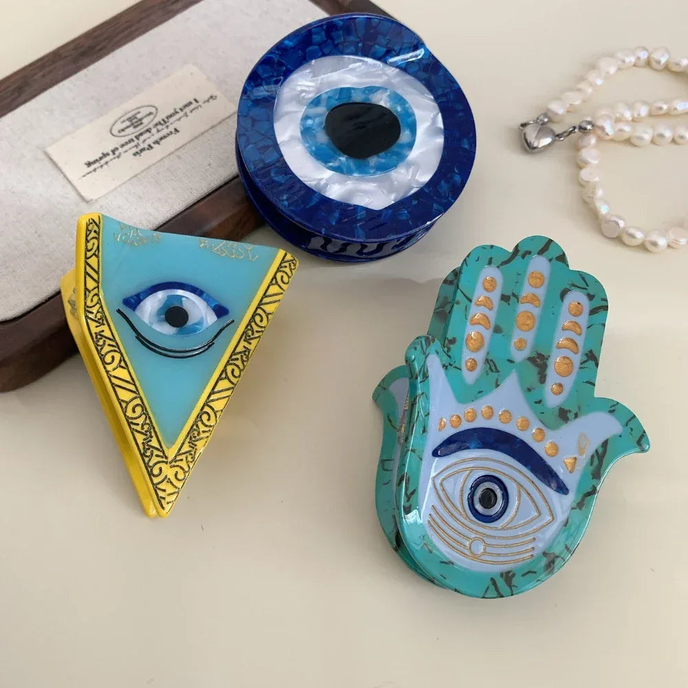 Three decorative accessories with evil eye motifs, including a Hamsa hand, a triangle, and a round disc, arranged with a string of pearls.