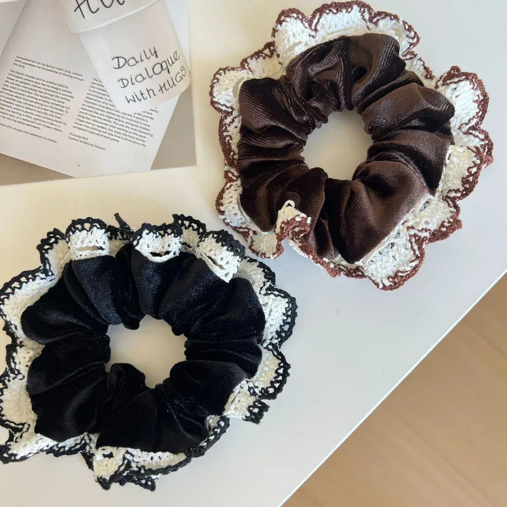 Two scrunchies, one brown and one black, featuring ruffled crocheted lace trim, are placed on a white surface with partially visible text and a brown box.