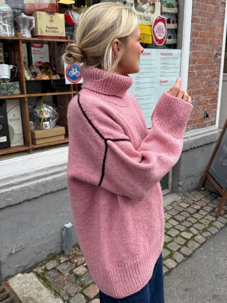 Woman in a pink oversized turtleneck sweater with contrasting black seam details and dark wash jeans, gesturing with her hand.
