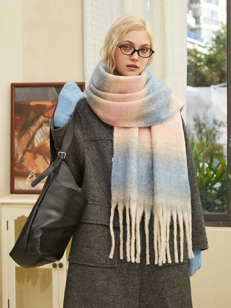 Close-up of a woman wearing a charcoal gray coat, light blue gloves, and a large ombre scarf with pink, blue, and cream colors. She is wearing round glasses.