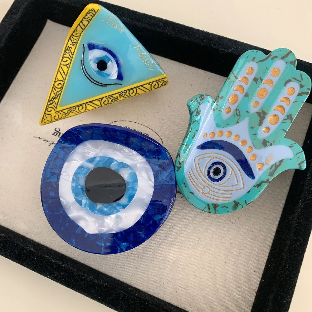 A collection of three evil eye themed accessories: a triangle, a round disc, and a Hamsa hand.