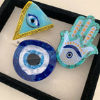 A collection of three evil eye themed accessories: a triangle, a round disc, and a Hamsa hand.