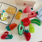 A close-up shot of fruit-shaped hair clips displayed on a white surface, with a tray and a box in the background.