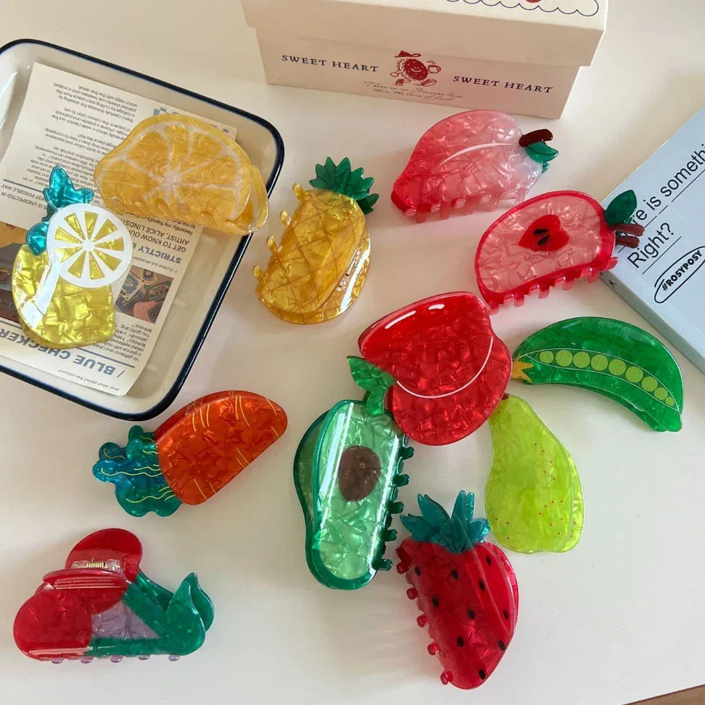 A close-up shot of fruit-shaped hair clips displayed on a white surface, with a tray and a box in the background.