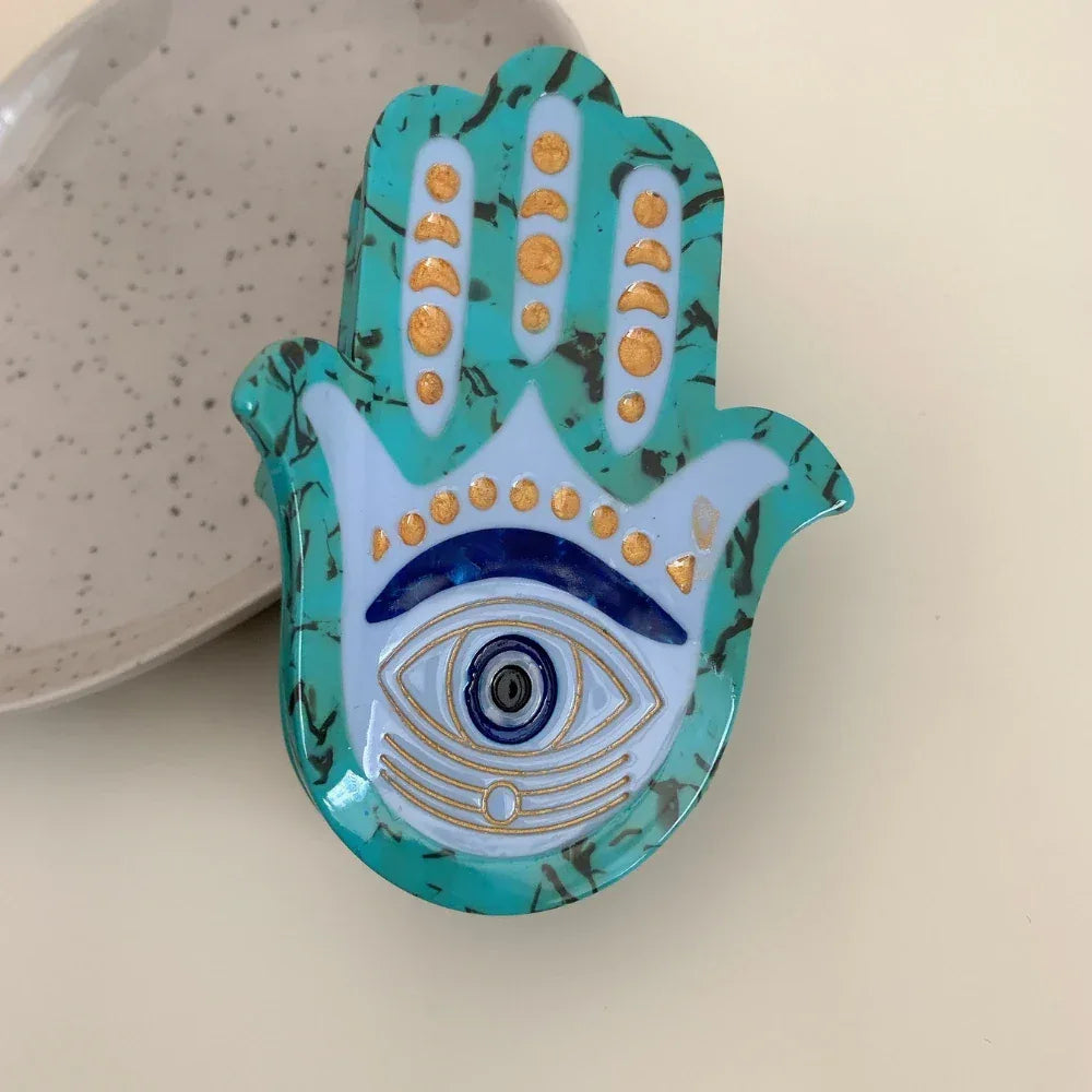 A turquoise Hamsa hand accessory with an evil eye motif, placed on a light-colored surface.