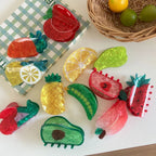 A flat lay of various fruit-shaped hair clips arranged on a white surface with a checkered cloth and a basket of fruit in the background.