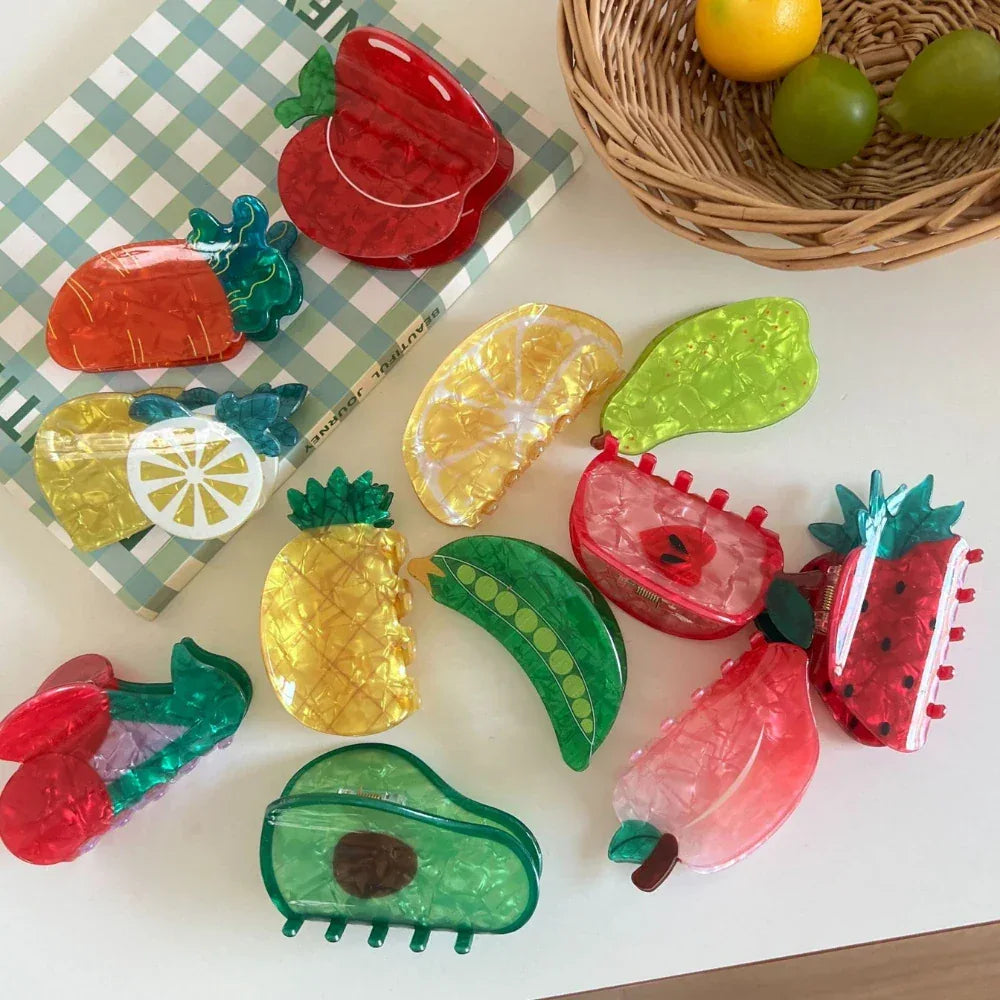A flat lay of various fruit-shaped hair clips arranged on a white surface with a checkered cloth and a basket of fruit in the background.