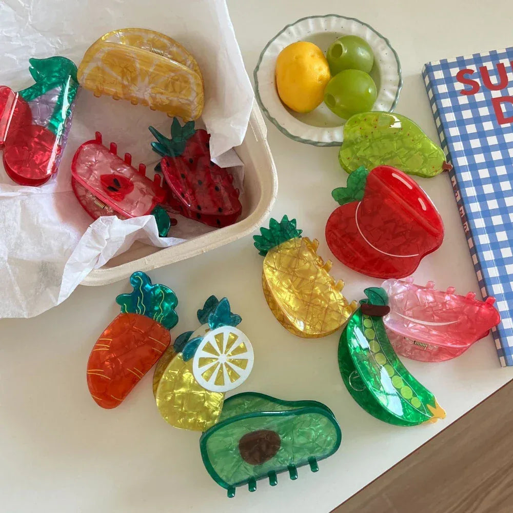 An overhead view of fruit-shaped hair clips scattered on a white table, with a box and a bowl of fruit visible.