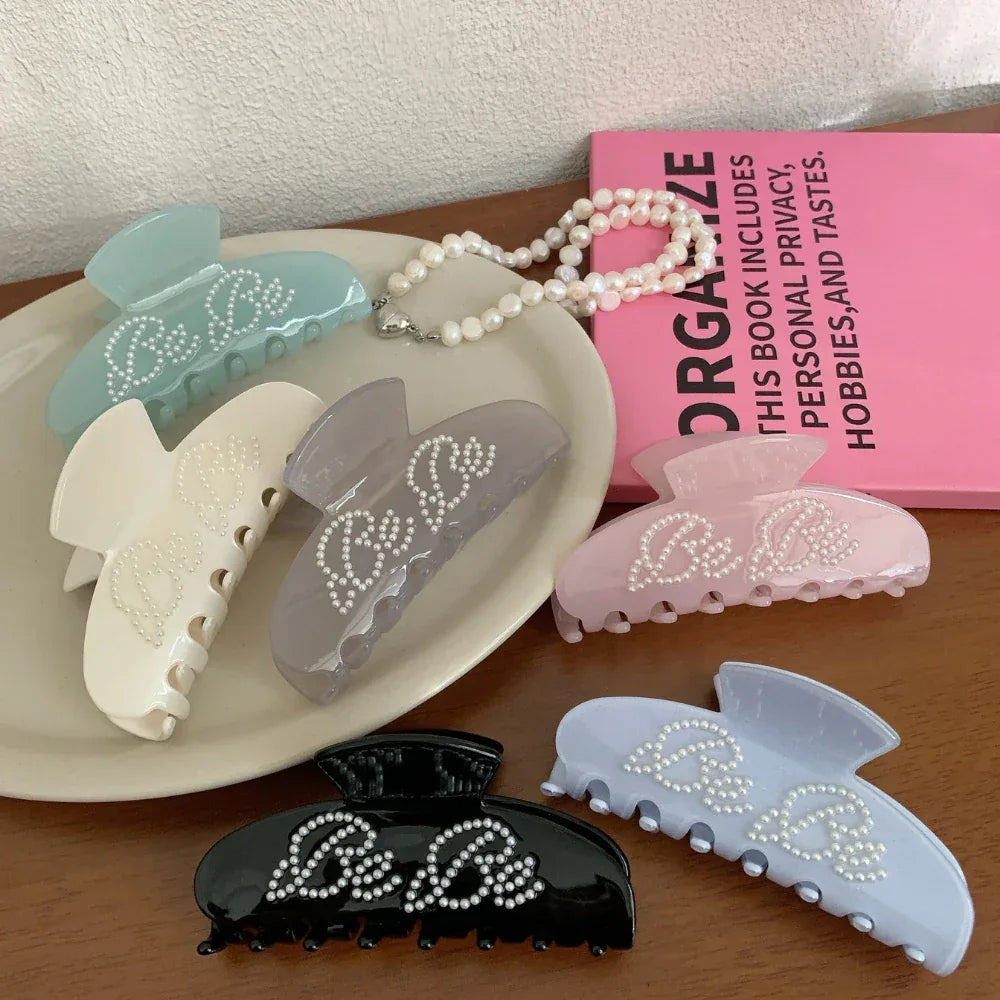 A group of pastel and dark-colored claw clips with the "Be Be" design, placed on a table with a pink book and pearl necklace.