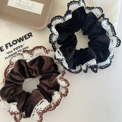 A top-down view of a brown scrunchie and a black scrunchie, both with decorative crocheted lace edges, are presented on a white surface next to a canvas tote bag.