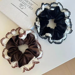 A brown scrunchie and a black scrunchie, each adorned with white and brown crocheted lace trim, are arranged on a white tabletop with a book in the background.