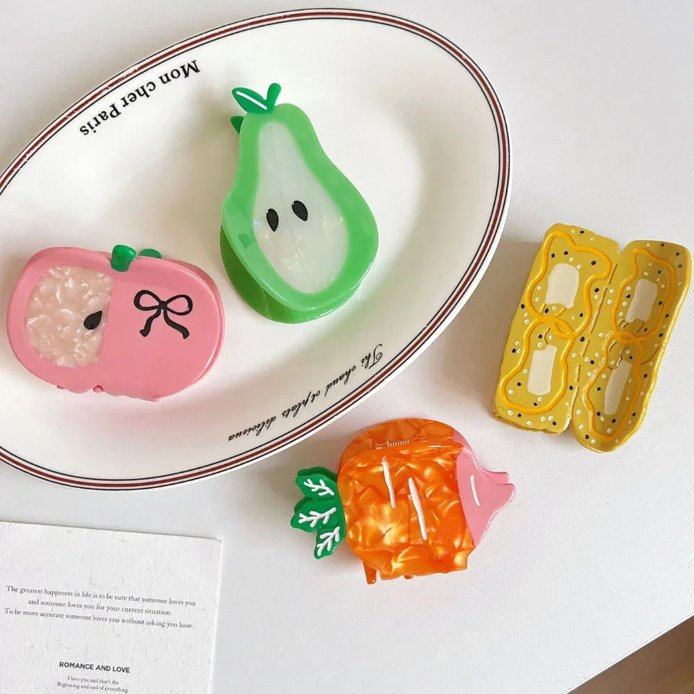 A white oval plate with text 'Mon cher Paris' holds a green pear hair clip and a pink apple hair clip. Next to it are a yellow banana slice hair clip and an orange carrot hair clip.