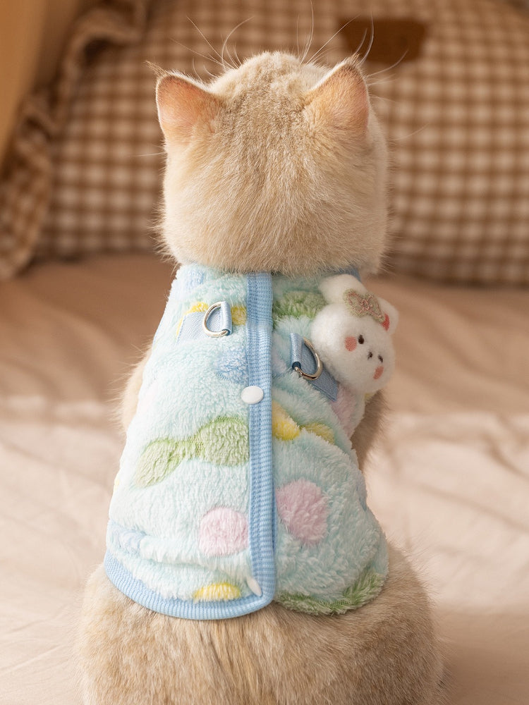 The back view of a cream-colored cat wearing a light blue fleece vest with a decorative charm.