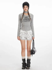 Model wearing a grey long-sleeved top with a graphic print and silver ruffled shorts, black beanie, white socks, and black boots.