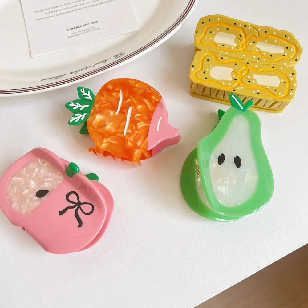 A top-down view of fruit-themed hair clips on a white surface: a pink apple, an orange carrot, a green pear, and a yellow banana slice design. A note and ribbon are also visible.