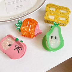 A top-down view of fruit-themed hair clips on a white surface: a pink apple, an orange carrot, a green pear, and a yellow banana slice design. A note and ribbon are also visible.