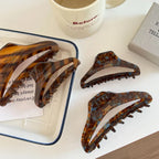 Several tortoiseshell claw hair clips are arranged on a white tray and a white table, with a mug and a book in the background.