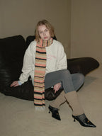 A woman sits on a black leather couch wearing a beige sweatshirt, gray leggings, leg warmers, and a striped scarf.