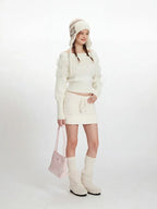 Model wearing a white off-the-shoulder sweater and matching mini skirt with a white knit hat, white knee-high socks, and white fuzzy boots. She is holding a pink handbag and posing sideways.