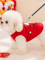 A white dog wearing a red corduroy vest with a leash attached, standing on a white surface.