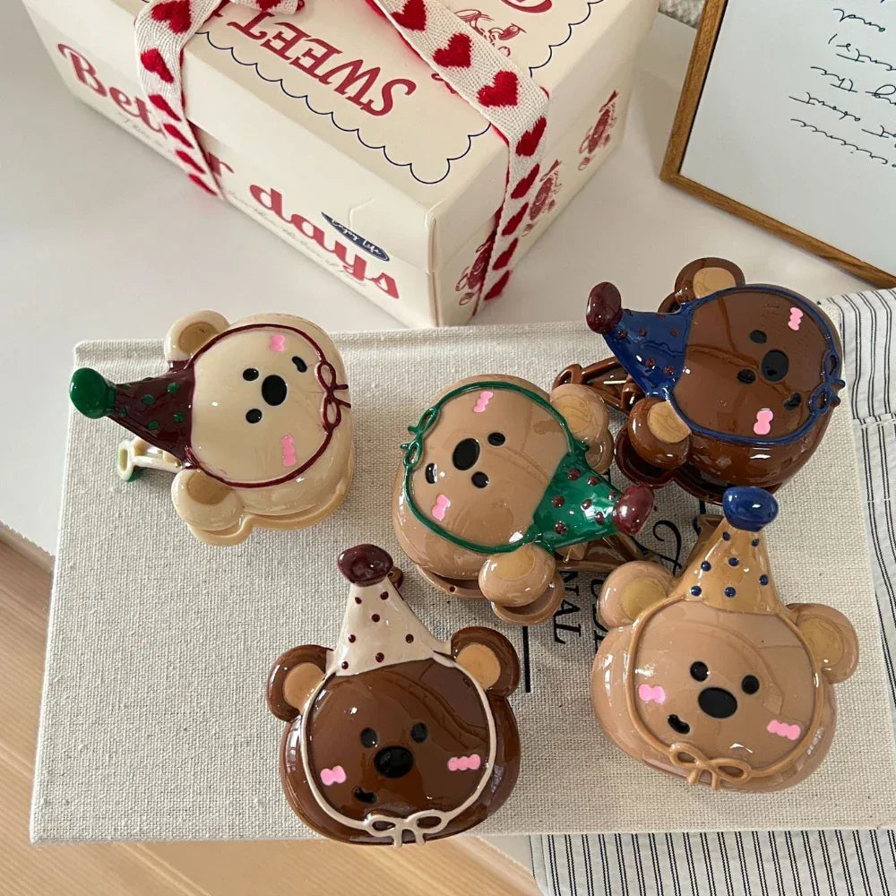 A collection of five bear-shaped hair clips with party hats of various colors, arranged on a textured surface with a gift box in the background.