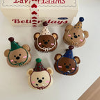 A top-down view of five bear-shaped hair clips with party hats, neatly arranged on a white surface, with a decorative gift box in the background.
