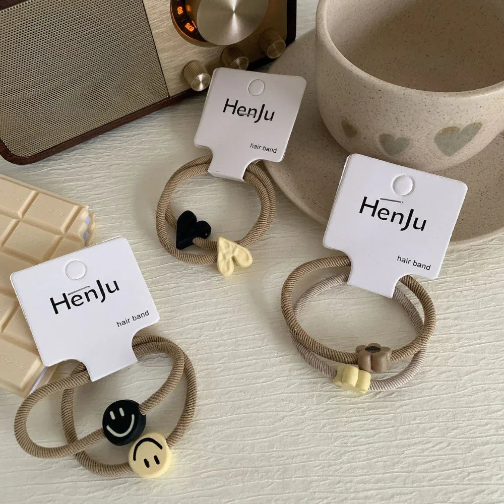 Three sets of beige hair ties with charms (smiley faces, hearts, flowers) are arranged on a table with a radio and cup.